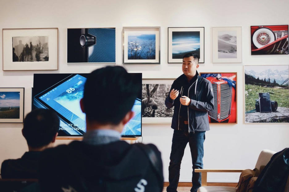 A man stands and speaks in front of a seated audience in a gallery-like room with framed photographs and artwork on the wall behind him. A large display screen shows a smartphone interface. The atmosphere suggests a presentation or workshop.